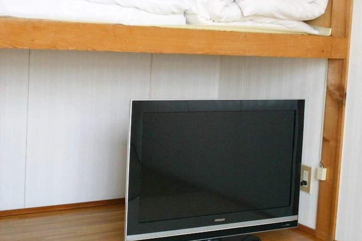 Una habitación con televisión sobre un soporte, un estante de madera con mantas dobladas y una pared blanca.