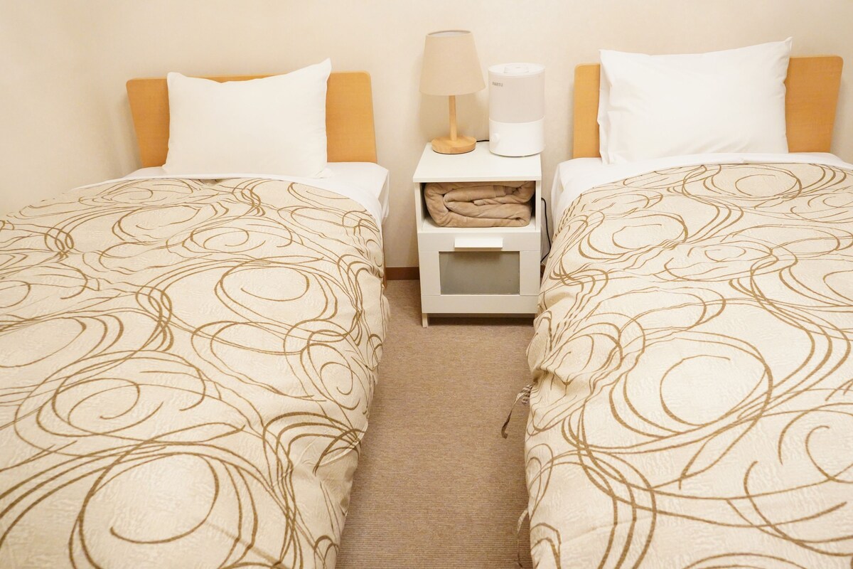 A hotel room with two single beds, a nightstand with a lamp and a device, and a small storage box.