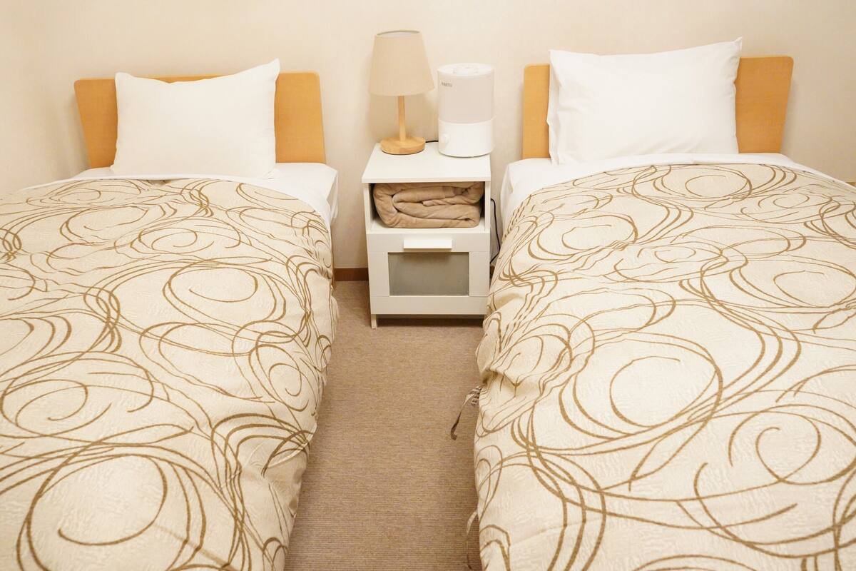 Quarto de hotel com duas camas de solteiro, mesa de cabeceira com uma lâmpada e um dispositivo, e uma pequena caixa de armazenamento.