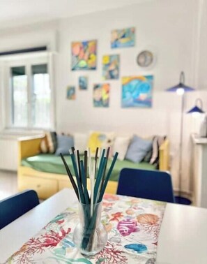 Une pièce comprenant un lit, une table sur laquelle se trouve un vase en verre, et un mur orné de tableaux colorés.
