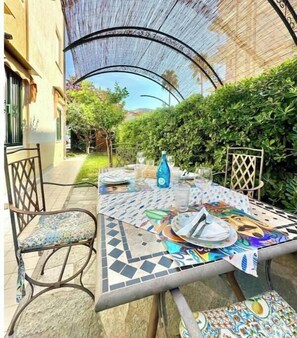 Area pranzo all'aperto con tavolo apparecchiato per due, circondato da sedie e un ombrellone decorativo.