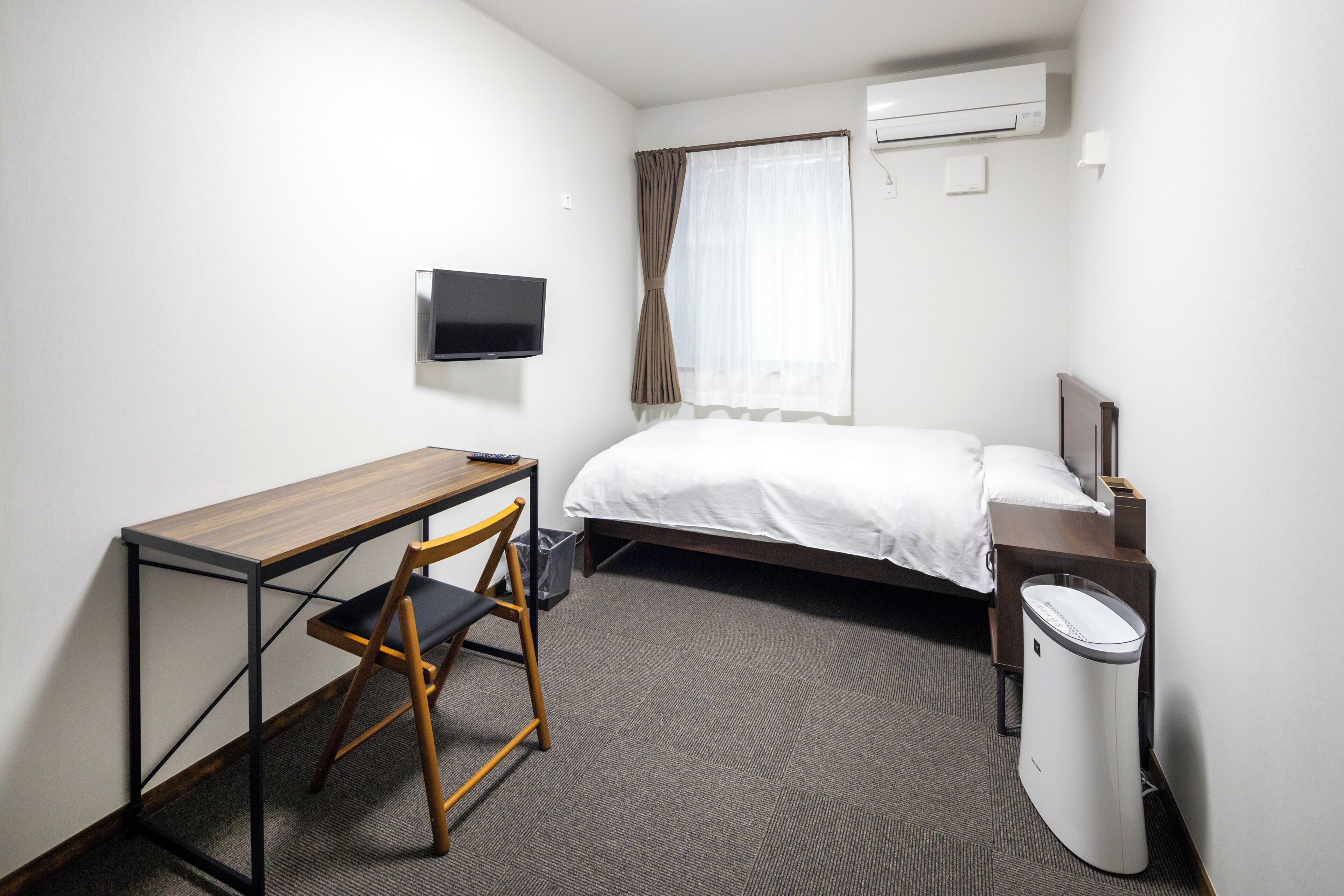 A hotel room with a bed, a desk with a chair, a TV, a waste bin, and an air conditioning unit.