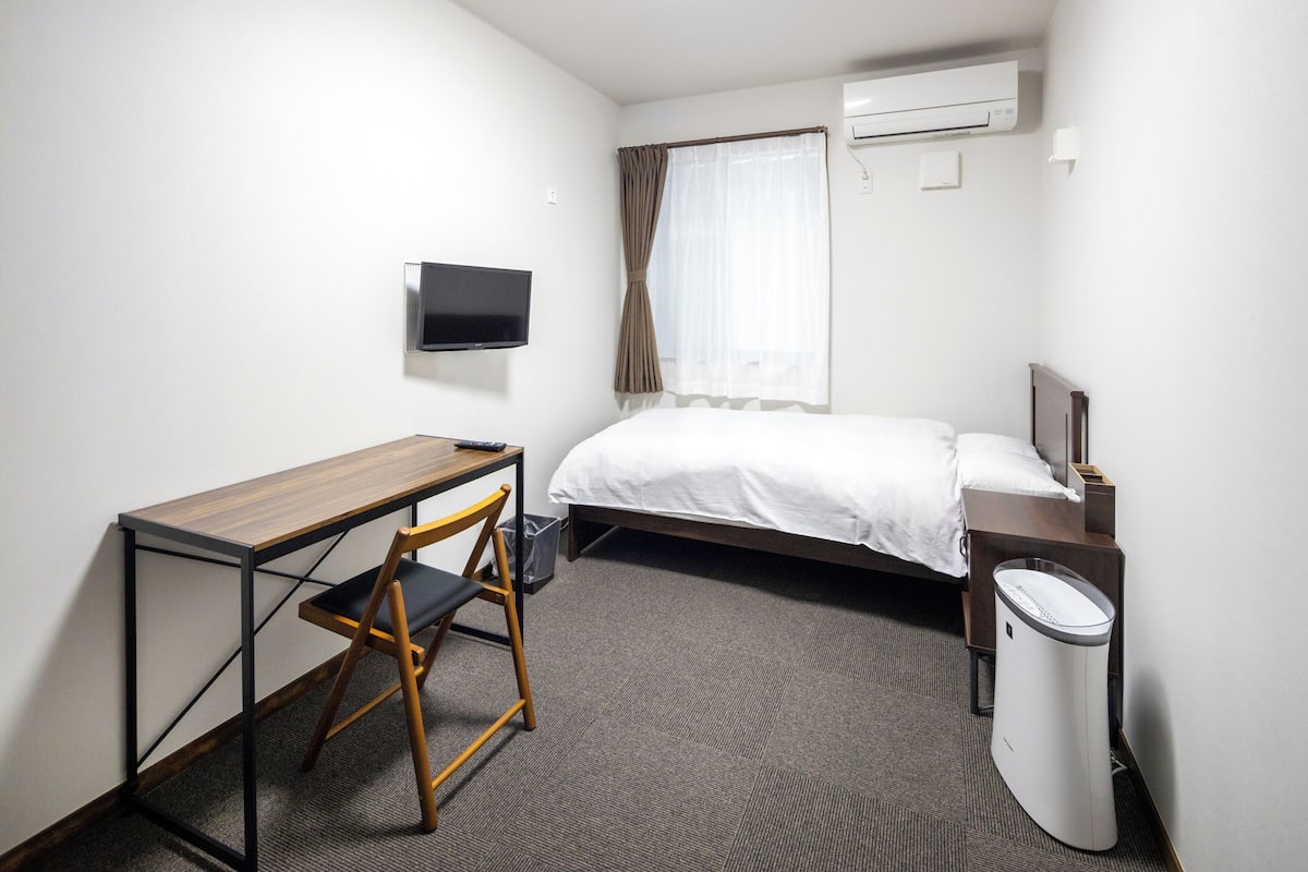 Ett hotellrum med en säng, ett skrivbord med en stol, en TV, en papperskorg och en luftkonditioneringsanläggning.