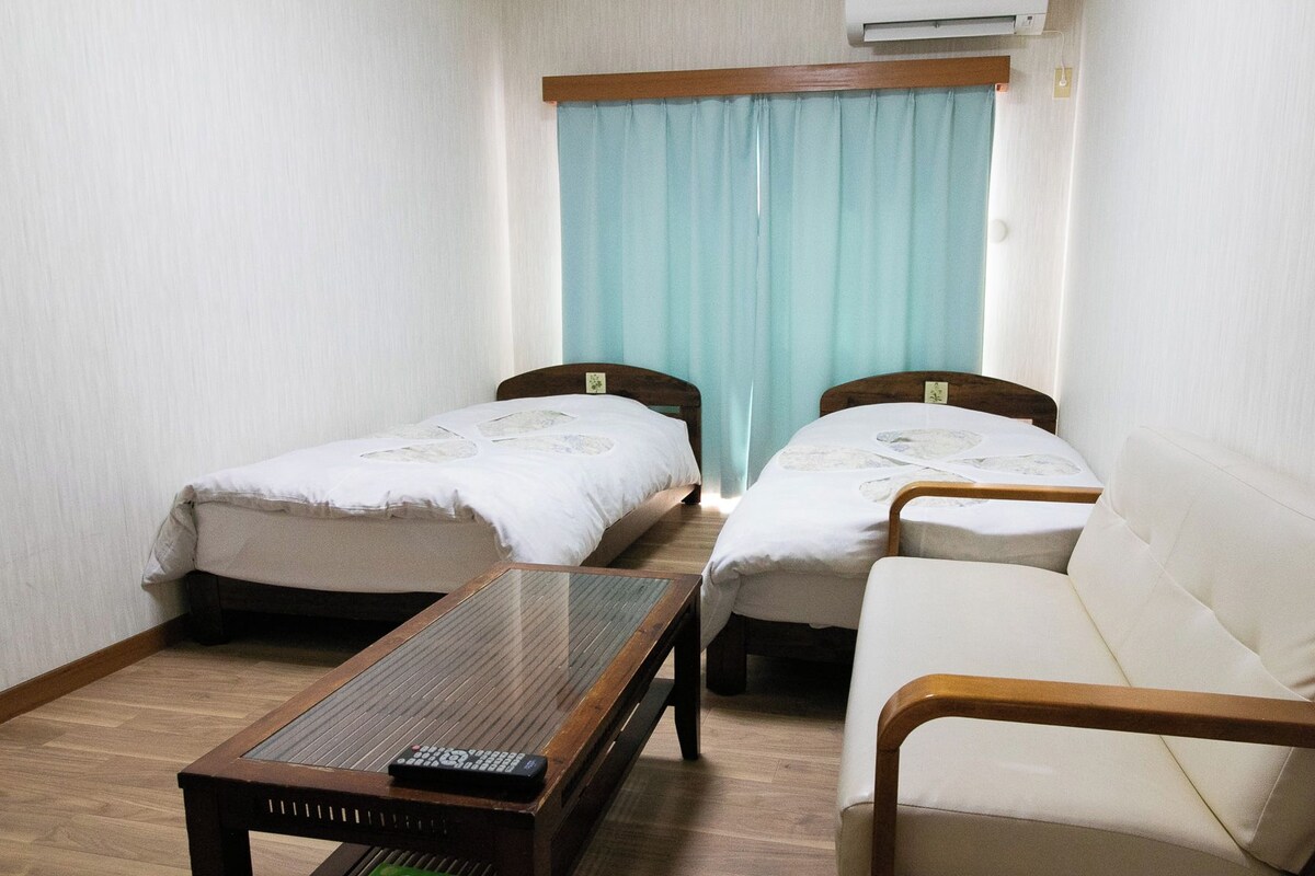 A hotel room with two single beds, a sofa, a wooden table with a remote control, and a window with curtains.