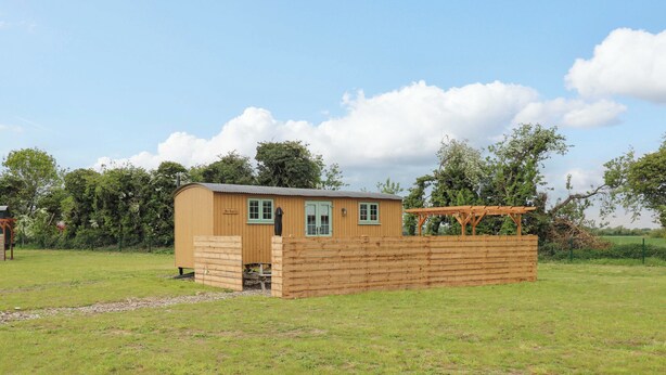 A small wooden structure with a door and windows, surrounded by a wooden fence, set in a grassy field with trees in the background. - The Kestrel (Cambridge)