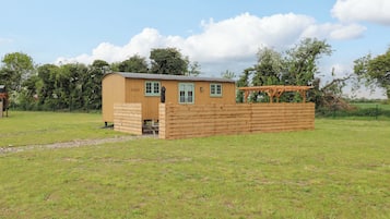 A small wooden structure with a door and windows, surrounded by a wooden fence, set in a grassy field with trees in the background.