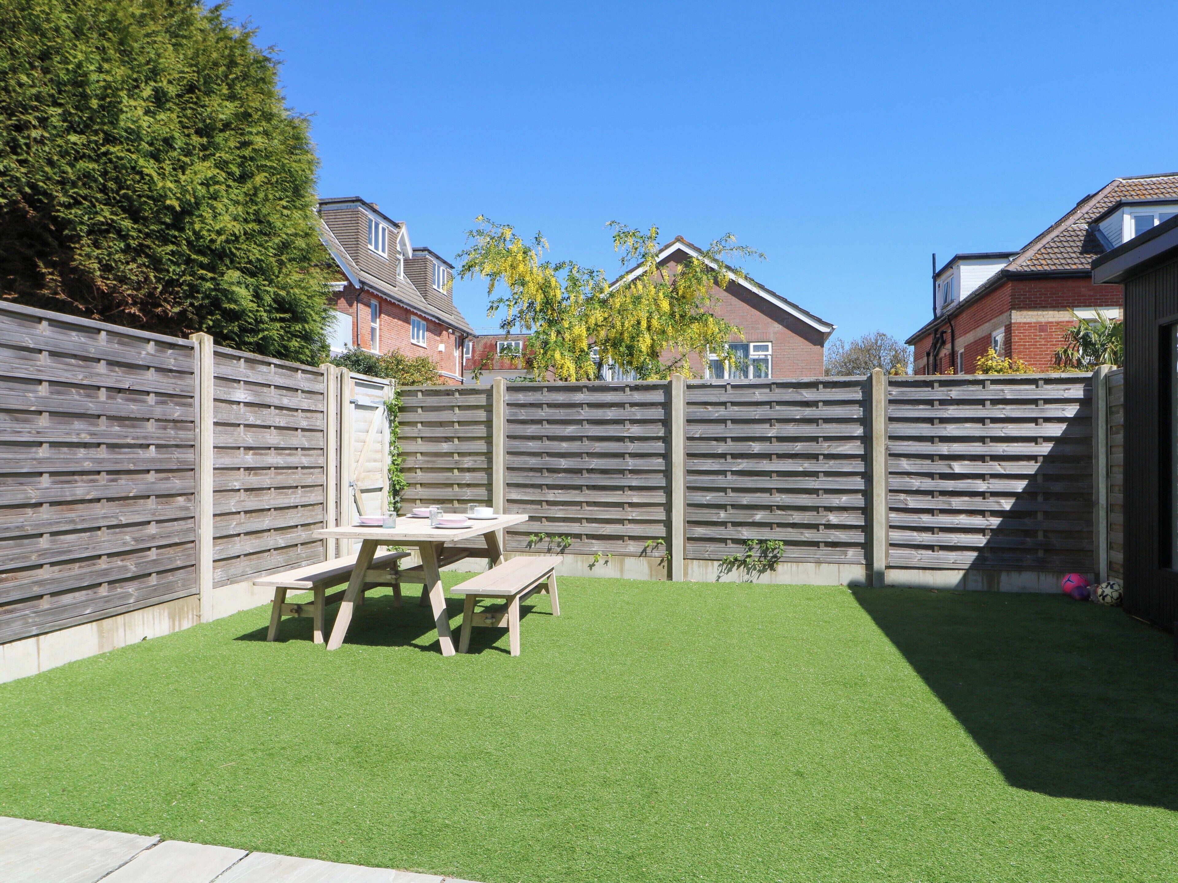 A backyard with a wooden fence, a picnic table, and artificial grass.