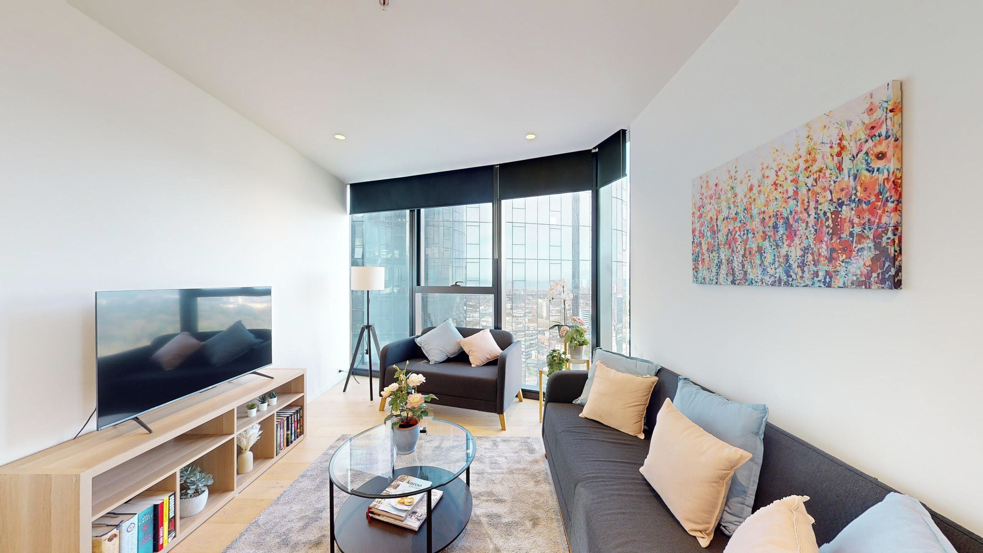 A modern living room with a TV, sofa, coffee table, and large windows offering a city view.