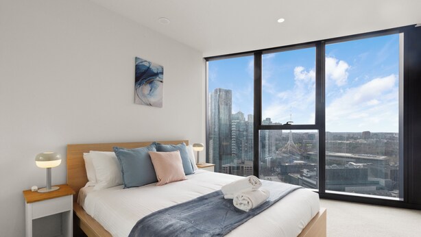 A modern bedroom with a large window, a bed with pillows, a nightstand, and a city view. - Melbourne Square Southbank (Southbank)