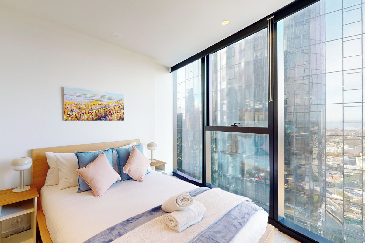 A modern hotel room with a large bed, a nightstand, a painting, and a city view through large windows.