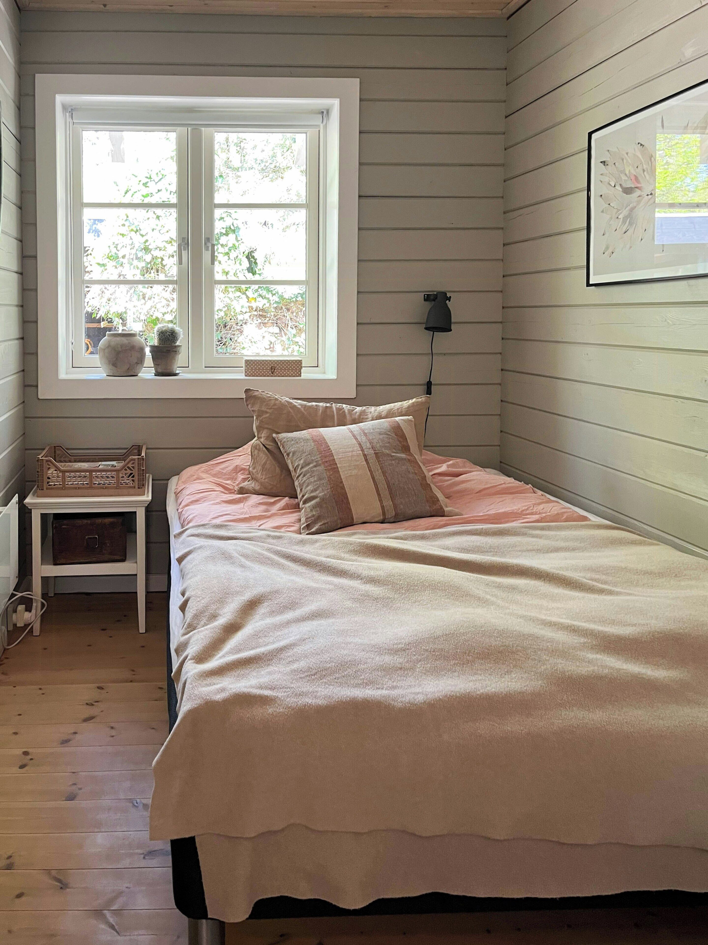 A bedroom with a bed, a nightstand, a lamp, a window with plants, and a picture on the wall.