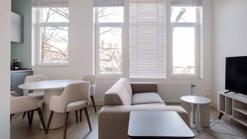 Een moderne woonkamer met een bank, eettafel, stoelen, een televisie en grote ramen.