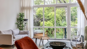 A modern living room with a large window, a sofa, a dining table, and a lamp. - Bright Ground-floor Apartment With Garden (Rotterdam)