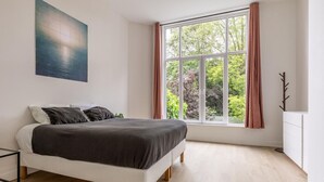 A bedroom with a bed, a window with curtains, a coat rack, and a painting on the wall. - Bright Ground-floor Apartment With Garden (Rotterdam)