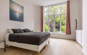 Ein Schlafzimmer mit einem Bett, einem Fenster mit Vorhängen, einem Kleiderständer und einem Gemälde an der Wand.