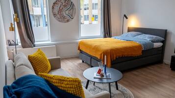 Een moderne hotelkamer met een bed, een bank, een rondend bijzettafeltje en uitzicht op de straat door grote ramen.
