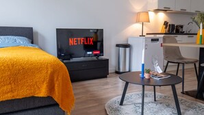 Ein modernes Hotelzimmer mit einem Bett, einem Fernseher, der 'NETFLIX' anzeigt, einem kleinen Tisch mit einer Lampe und einer Küchenzeile.