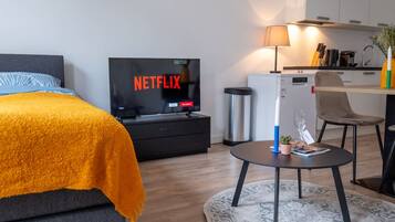 Een moderne hotelkamer met een bed, een televisie waarop 'NETFLIX' wordt weergegeven, een tafeltje met een lamp en een kitchenette.