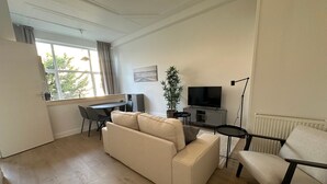 Uma sala de estar moderna com um sofá bege, uma mesa de jantar com cadeiras, uma TV, uma planta e um radiador.