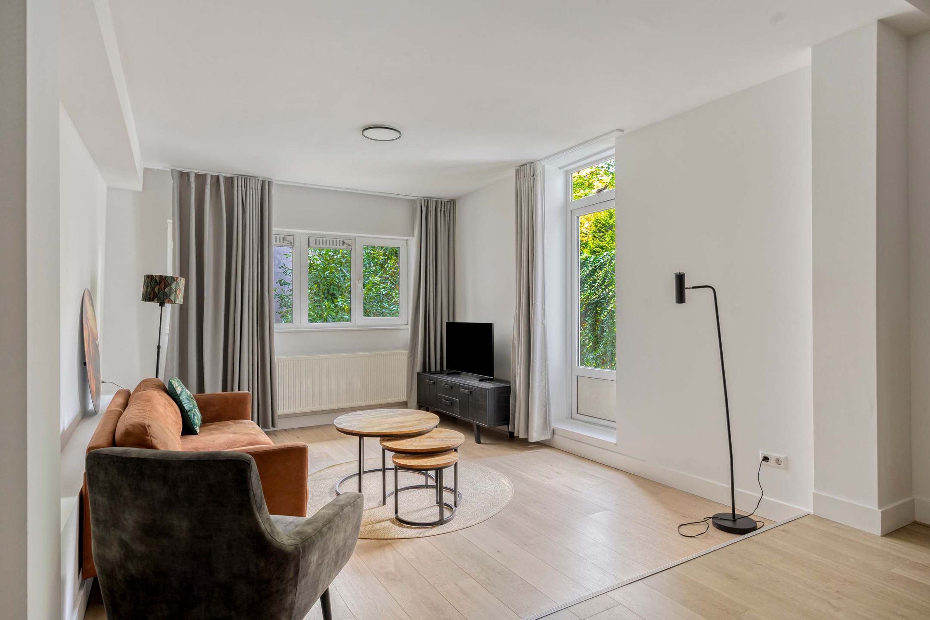 Una sala de estar moderna con un sofá, dos mesas redondas, un televisor y ventanas amplias con cortinas.