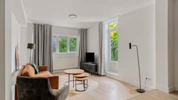 Una sala de estar moderna con un sofá, dos mesas redondas, un televisor y ventanas amplias con cortinas.