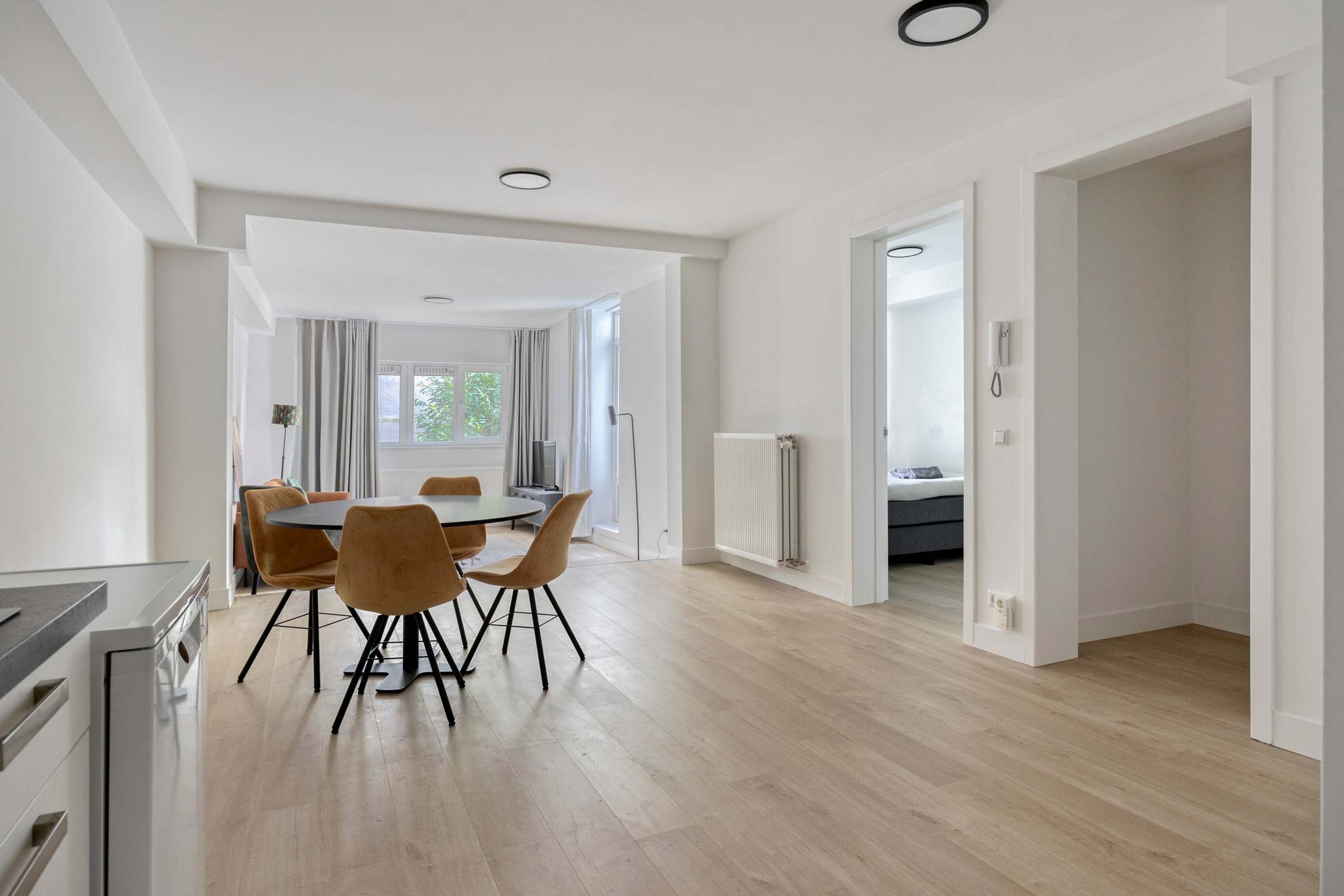 A modern kitchen with a built-in oven, a dining area with a round table and chairs, and a view of a bedroom through an open door.