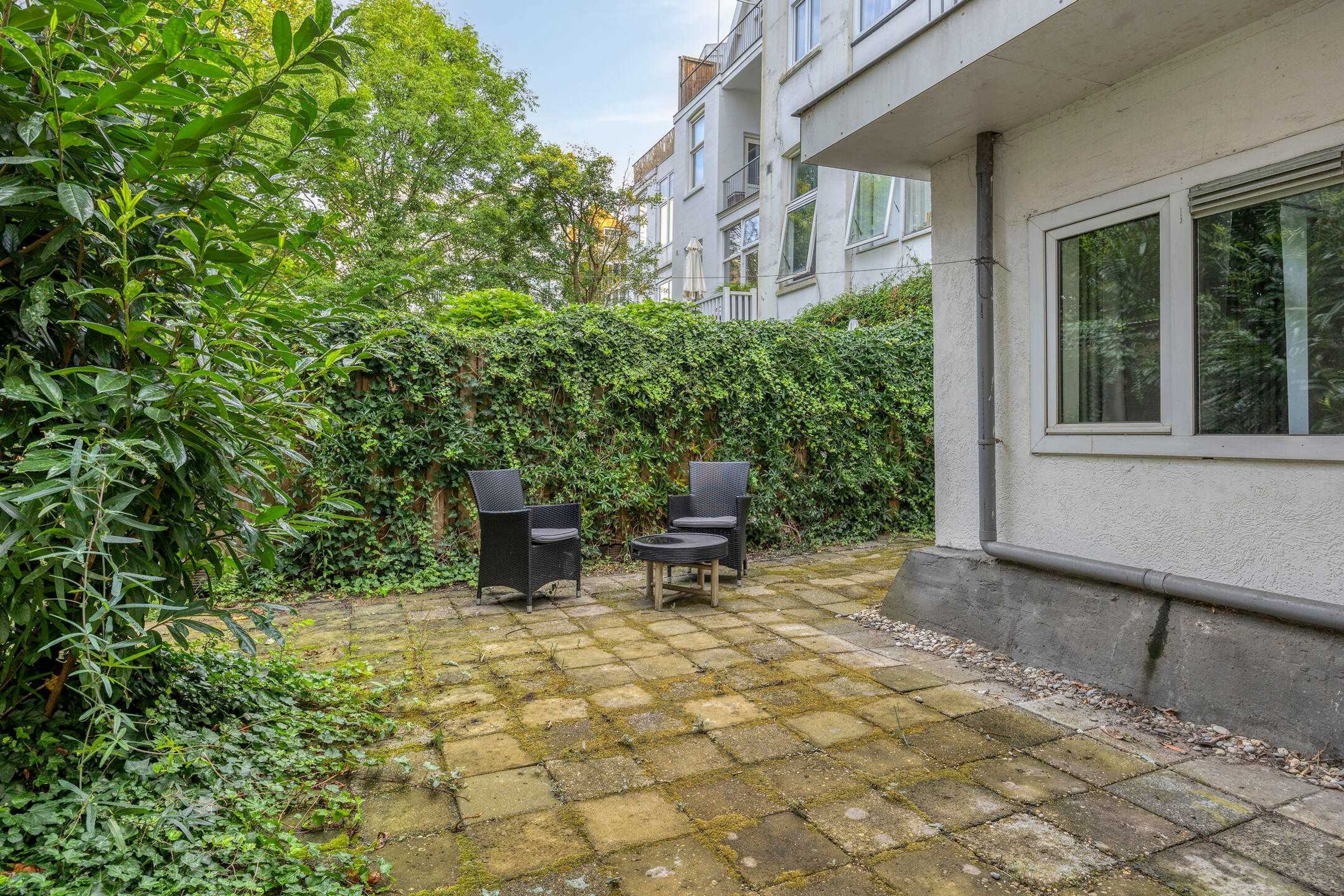 Un patio con suelo empedrado, dos sillas de mimbre y abundante vegetación.