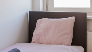 Una cama individual con cabecera, una ventana con persianas y una toalla enrollada sobre la cama.