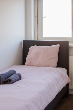 Ein Einzelbett mit Kopfteil, ein Fenster mit Jalousien und ein aufgerolltes Handtuch auf dem Bett.