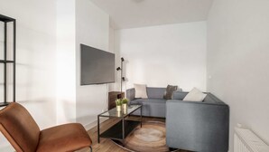 Un salón moderno con un sofá, una mesa de centro de cristal, un sillón marrón y un televisor instalado en la pared.