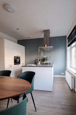 A modern kitchen with white cabinets, a central island, and a dining area with a wooden table and green chairs. - Modern 2BR Apartment Near Rotterdam Central Station (Rotterdam)