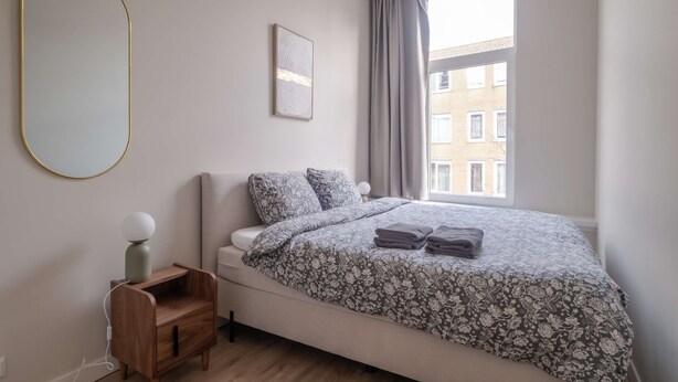 Un dormitorio con cama, mesita de noche con lámpara, ventana con cortinas y espejo montado en la pared.