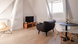 Et loftrum med TV, spisebord med stole, en fletstol og en potteplante.
