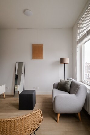 A modern living room with a grey sofa, a black ottoman, a wicker chair, a floor lamp, a mirror, and a framed picture on the wall. - Cosy City Apartment in Rotterdam West (Rotterdam)