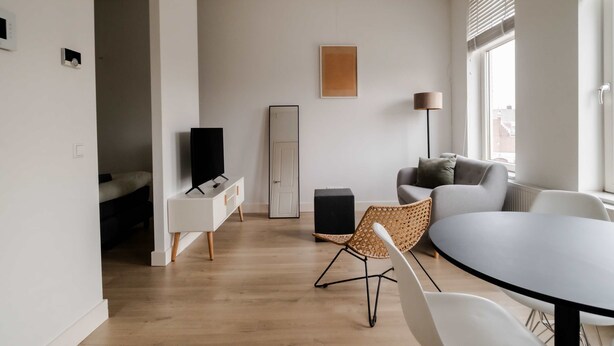 A modern living room with a flat-screen TV, a sofa, a dining table with chairs, and a large window with blinds. - Cosy City Apartment in Rotterdam West (Rotterdam)