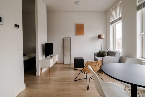 Una sala de estar moderna con un televisor de pantalla plana, un sofá, una mesa de comedor con sillas y un ventanal con persianas.