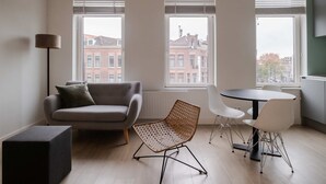 A modern living room with a sofa, a wicker chair, a dining table with chairs, and large windows with blinds. - Cosy City Apartment in Rotterdam West (Rotterdam)