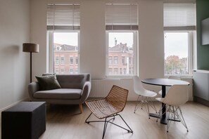 Una sala de estar moderna con un sofá, una silla de mimbre, una mesa de comedor con sillas y ventanas grandes con persianas.