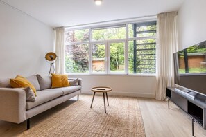 A living room with a sofa, a small round table, a television, and a large window with curtains. - Modern Ground-floor Apartment With Garden (Rotterdam)