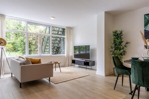 Ein modernes Wohnzimmer mit Sofa, Fernseher, Esstisch, Stühlen und einem großen Fenster mit Blick auf Bäume.