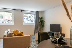 Uma sala de estar moderna com um sofá, uma mesa de jantar com uma taça de vinho, uma televisão e vista para a rua através de grandes janelas.