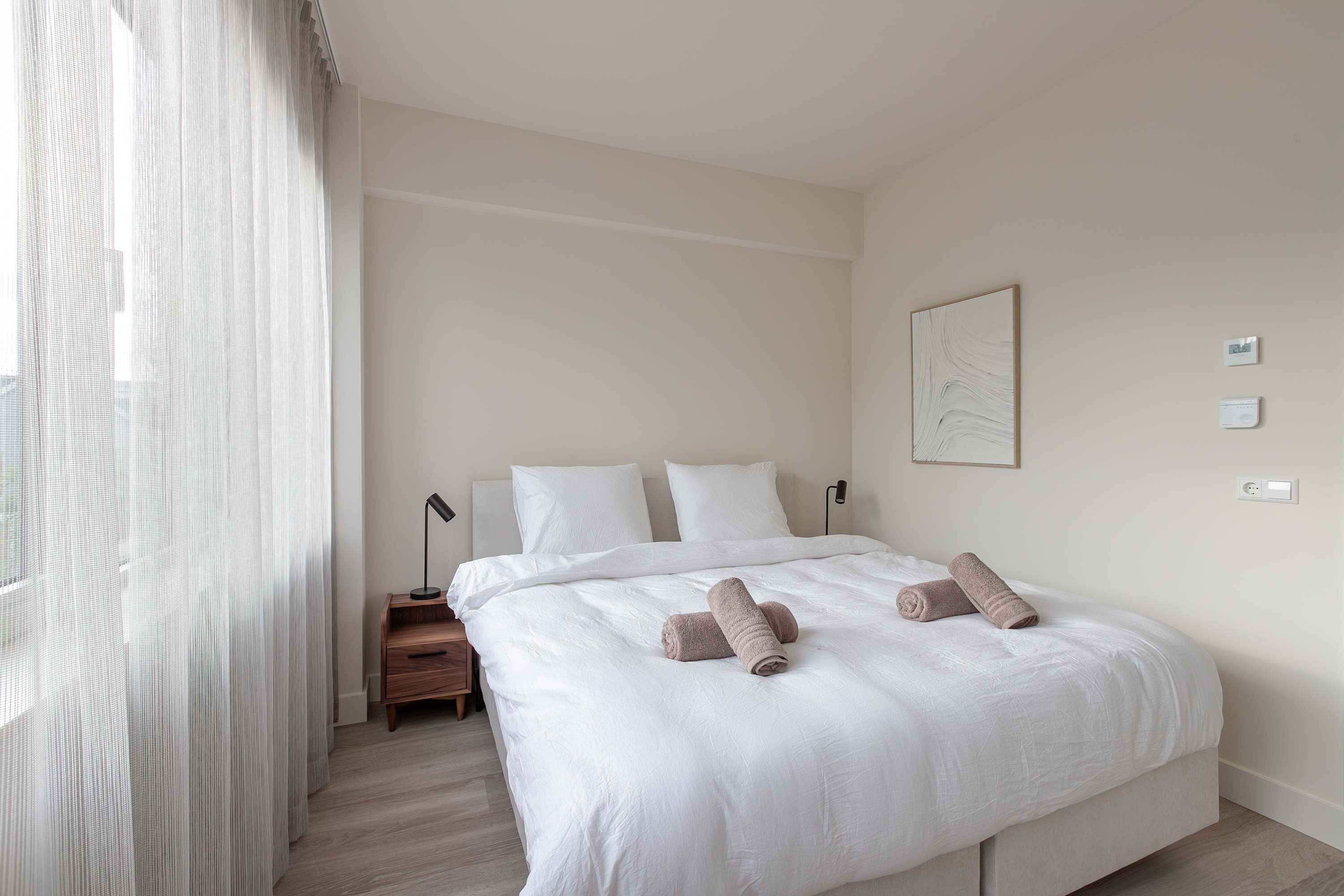 A neatly made bed with white linens, a bedside table with a lamp, and a window with sheer curtains.
