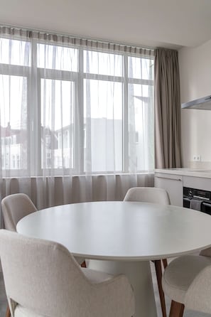 A modern room with a round table, six chairs, a kitchen area, and a large window with sheer curtains.