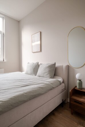 Ein Schlafzimmer mit einem Bett, einem Nachttisch, einem Spiegel und einem Fenster.