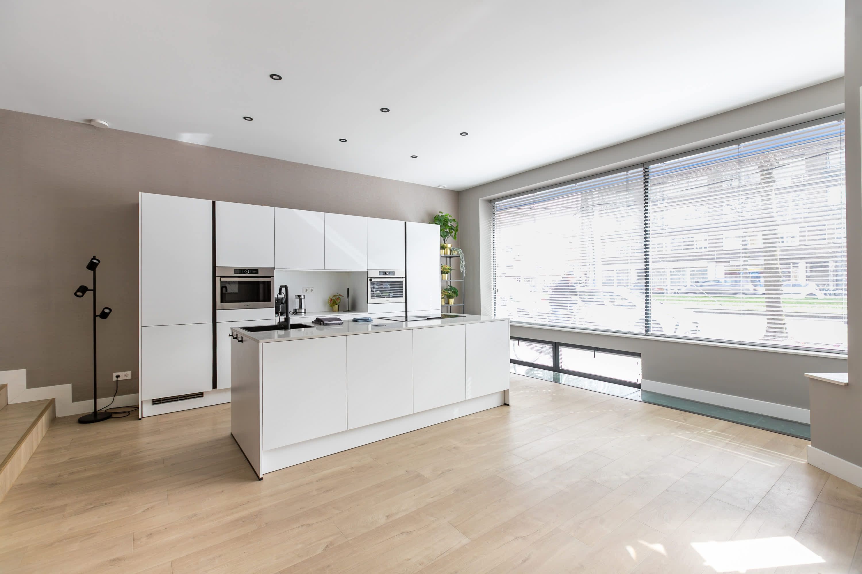 Een moderne keuken met witte kastjes, een centraal eiland en een groot raam met zonwering.