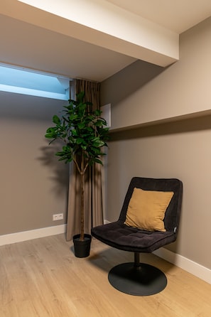 Ein Zimmer mit einem Blumentopf, einem schwarzen Stuhl mit gelber Kissenauflage, Holzdielen und einem Fenster mit Vorhängen.