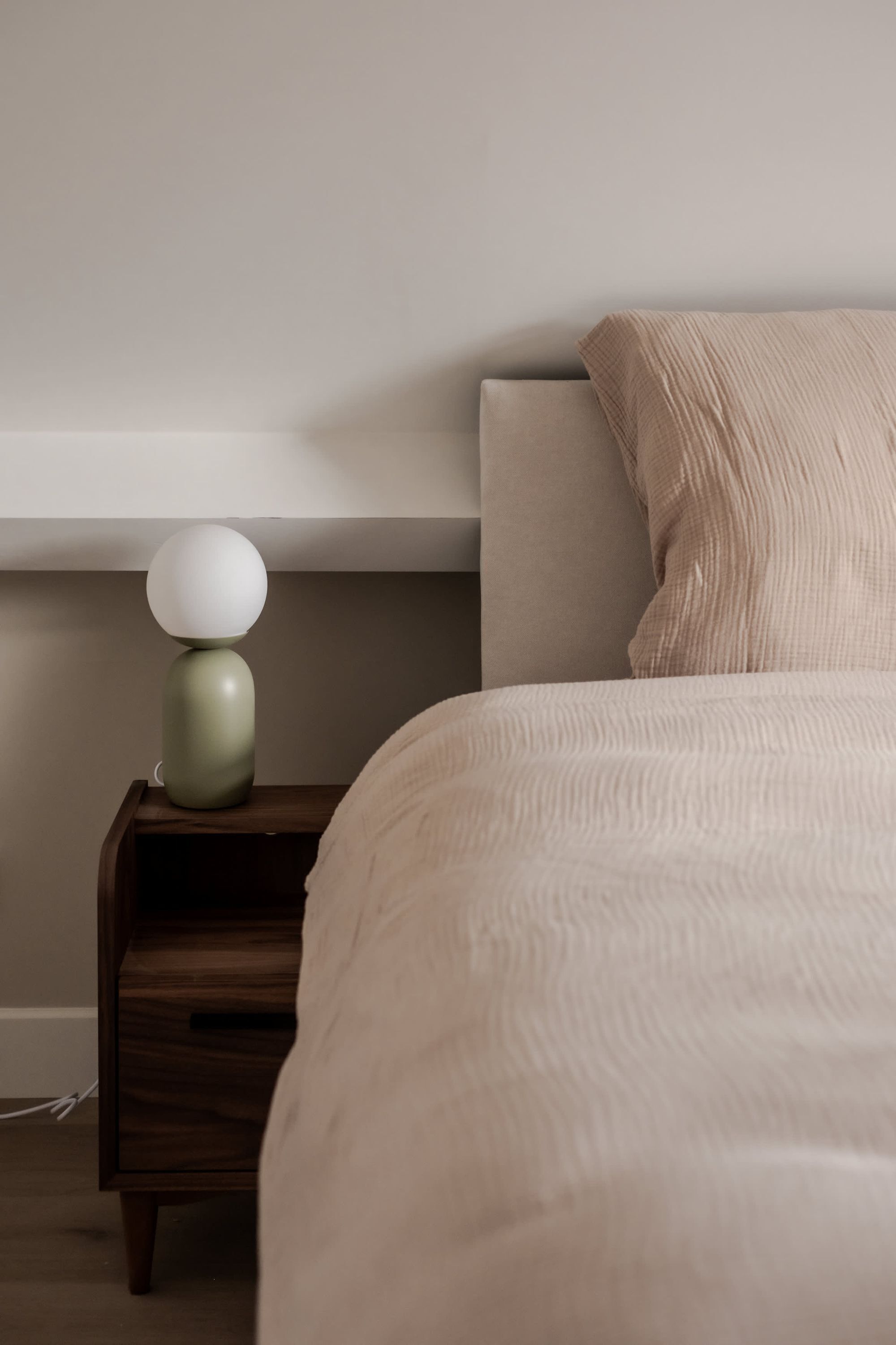 A bedroom with a bed, a nightstand with a lamp, and a wall-mounted shelf.