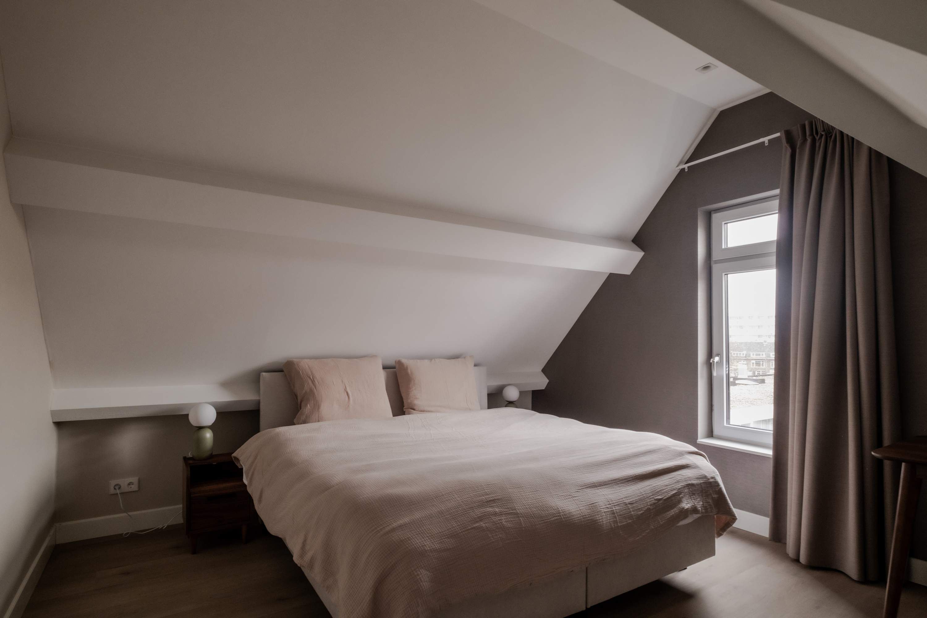A bedroom with a sloped ceiling, a bed, a nightstand, a lamp, and a window with curtains.