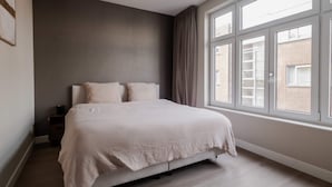 Ein Schlafzimmer mit großem Fenster, einem Bett mit weißer Bettdecke, einem Nachttisch und einer Wand mit strukturierter Gestaltung.
