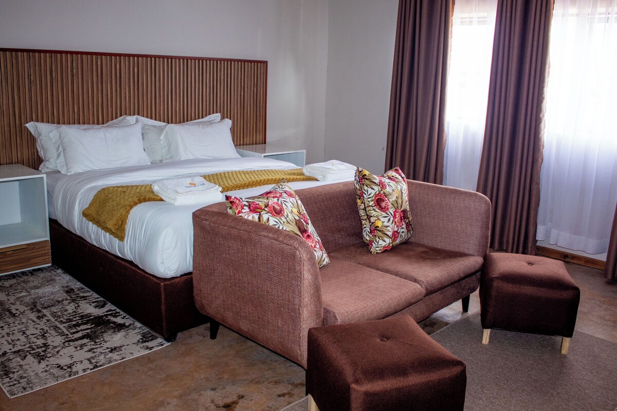 A bedroom with a bed, a sofa, an ottoman, a nightstand, and a window with curtains.